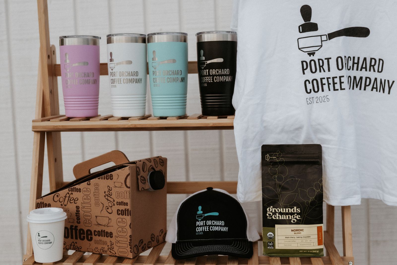 Merch shelf with tumblers in pink, white, teal and black, a trucker hat, a t-shirt, a coffee carafe box and a bag of beans