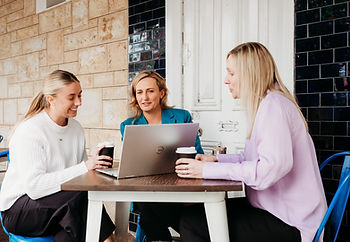 Three women sit at a table outdoors, with one using a laptop and all holding coffee cups. The mood is focused and collaborative.