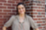 Shauna smiling, leaning against a brick wall. She wears a patterned blouse and necklace, with curly hair and hoop earrings, exuding warmth.