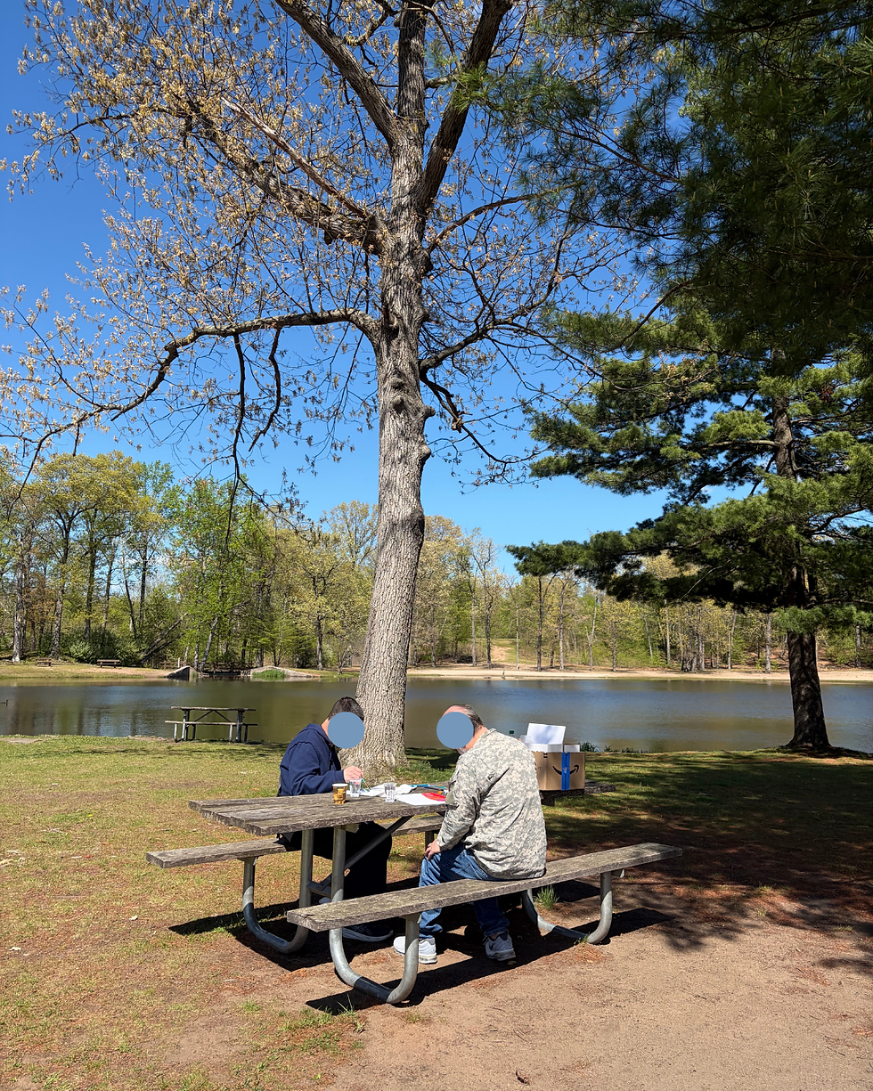 Two people sit at a picnic table by a lakeside under a tall tree, chatting and working on papers on a sunny spring day.