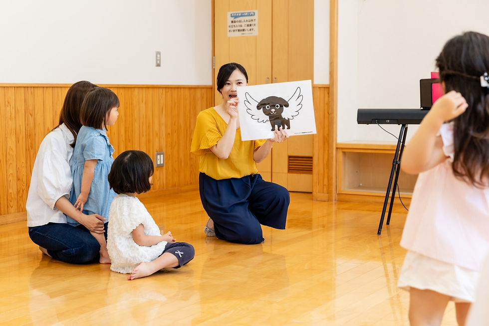 室内で女性がイラストを見せ、子どもたちが膝をついて見つめる。明るい木床の教室、和やかな雰囲気