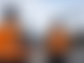 Two FAA-certified pilots in orange vests operate drones over a construction site with cranes and buildings. Clear blue sky above.