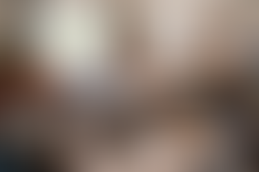 A messy bedroom with scattered clothes, books, and pizza box. Bed with gray sheets, guitar, plants, laptop on desk, and a corkboard on wall.