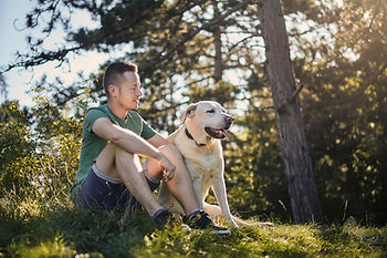 Man sitting on grass beside tan dog with trees in the background