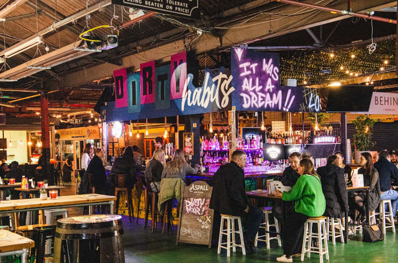 Crowded indoor bar with people at stools, neon graffiti signs reading Dirty Habits and It Was All a Dream, warm lively atmosphere