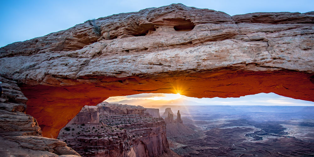 A sun-drenched view through Mesa Arch in Canyonlands National Park at sunrise, showing dramatic red rock canyons and distant blue mountains.