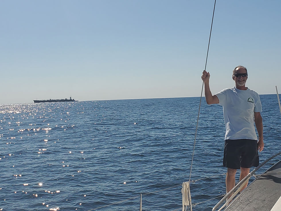 Boat owner Tom on his sailboat , on the ocean with a large cargo ship in the background