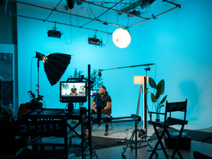 Video studio with blue backdrop, cameras and lights, a seated man being filmed, and Myers Media chairs in foreground.