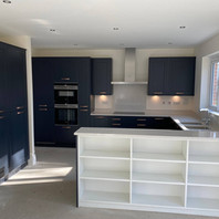 Modern kitchen with dark blue cabinets, built-in oven and integrated washing machine, and a white island featuring open shelving. Light countertops and a stainless steel range hood complete the bright, minimalist space with large windows.