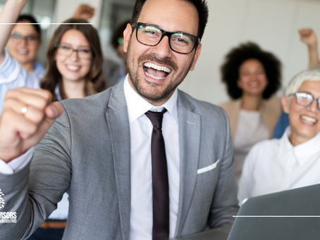 Group of people in an office, smiling and raising fists in celebration. Man in gray suit in foreground holds a laptop. Bright, joyful mood.