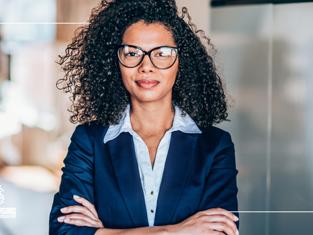 Confident woman with curly hair and glasses in a blue suit, arms crossed, stands in an office. TBG Advisors logo is visible.