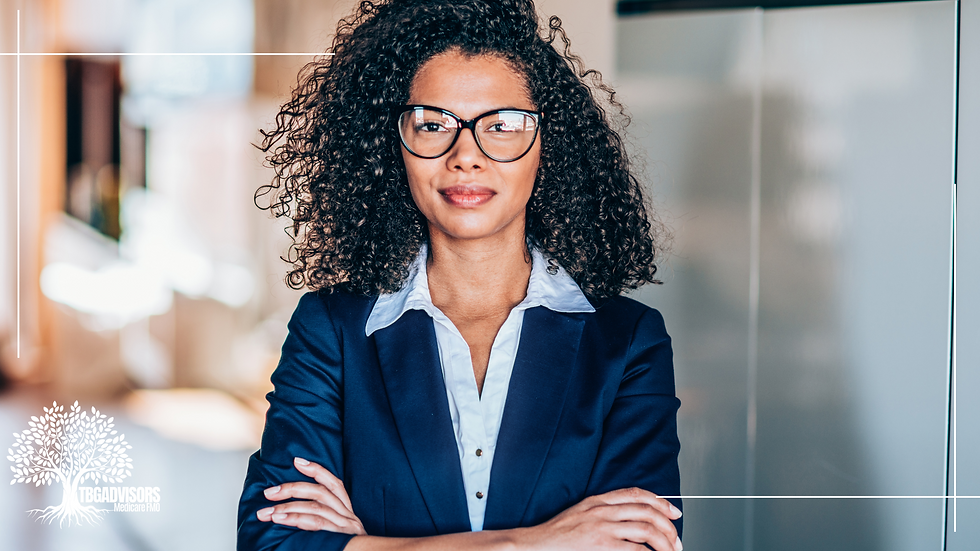 Confident woman with curly hair and glasses in a blue suit, arms crossed, stands in an office. TBG Advisors logo is visible.