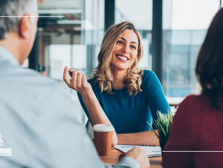 Smiling woman in blue shirt talks with two people in an office. Coffee cup and papers on table. Logo: