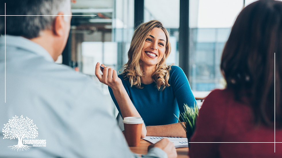 Smiling woman in blue shirt talks with two people in an office. Coffee cup and papers on table. Logo: