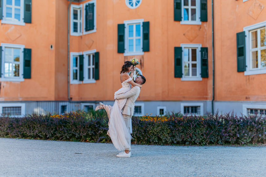 Bräutigam hebt Braut hoch, vor einem orangefarbenen Gebäude, Glückliche Hochzeit