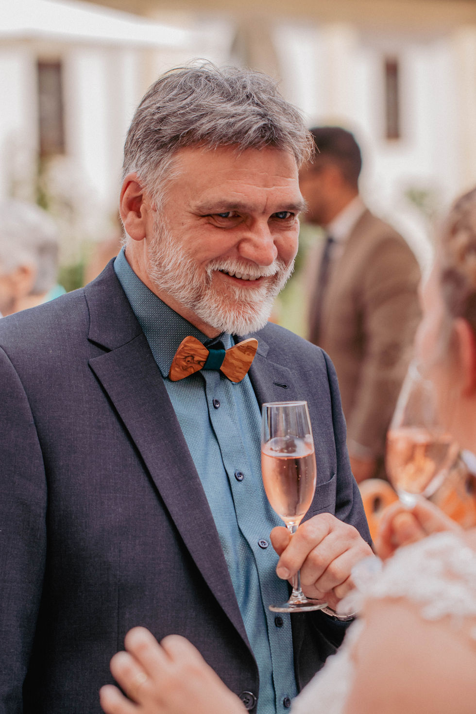 Ein Mann mit Holzfliege hält ein Glas Sekt, Jasmin & Oliver, Hochzeitsfeier.