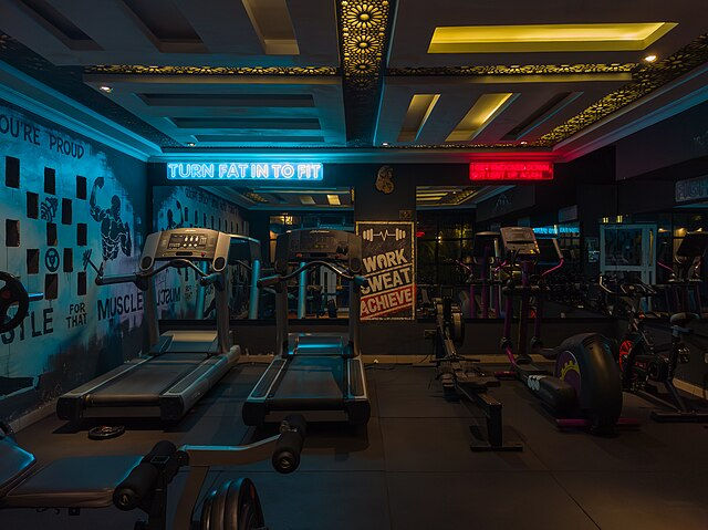 Dimly lit gym with treadmills and exercise bikes. Neon signs display "TURN FAT INTO FIT" and "WORK, SWEAT, ACHIEVE" on the walls.