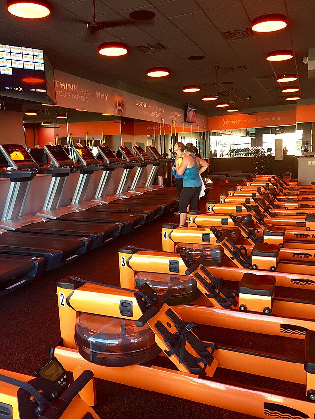 Gym with orange rowing machines and treadmills. Two people are using equipment. Motivational text on walls, warm lighting.