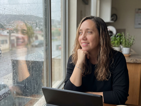 A young woman with wavy brown hair, a warm smile, and an eye bracelet sits at a wooden table, resting her chin on her hand while gazing thoughtfully out a large, rain-streaked window. She is at her home kitchen table with a Microsoft Surface Pro laptop open and a cup of latte art coffee. The outside view is a soft, blurry landscape of a CIty in Mexico, during the rain. Her reflection is visible on the wet glass.