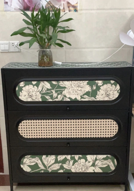 Black dresser with floral and woven patterns, potted plant on top. Light beige tiled wall background. Cozy and decorative setting.