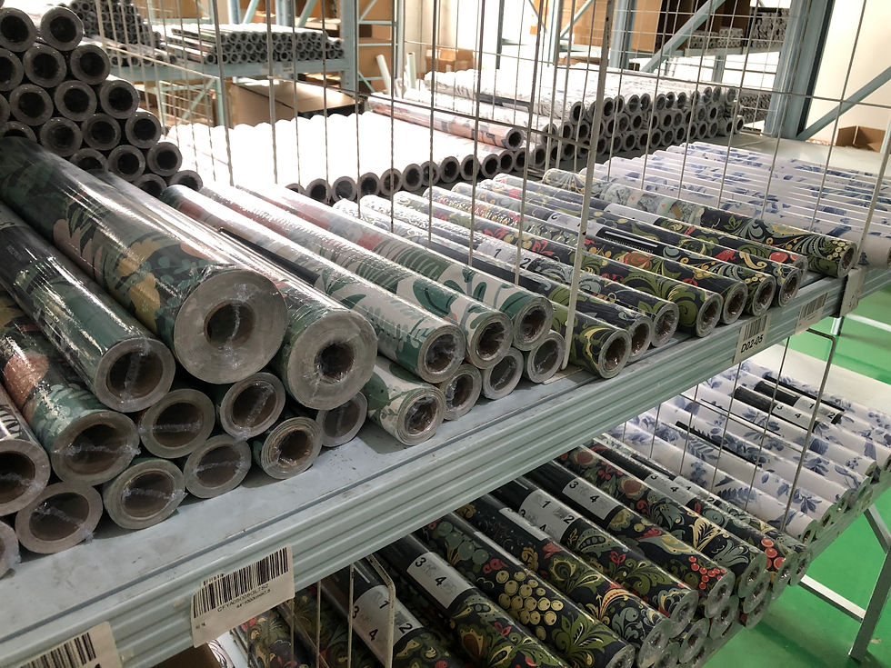 Rolls of colorful patterned wallpaper are stacked on metal shelves in a store. Background shows more shelves with organized patterns.