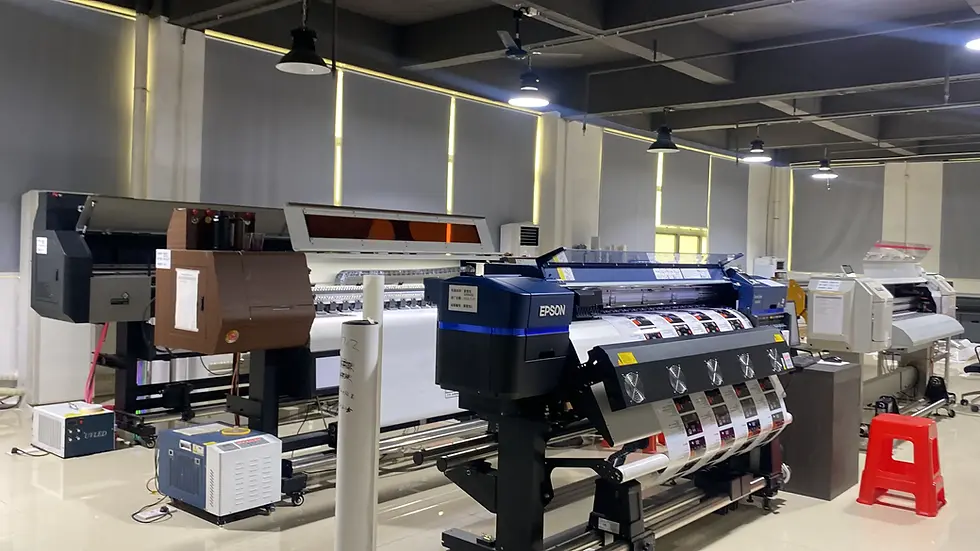 Printing machines in a well-lit room with gray walls. An Epson printer is prominent, surrounded by various equipment. No people present.