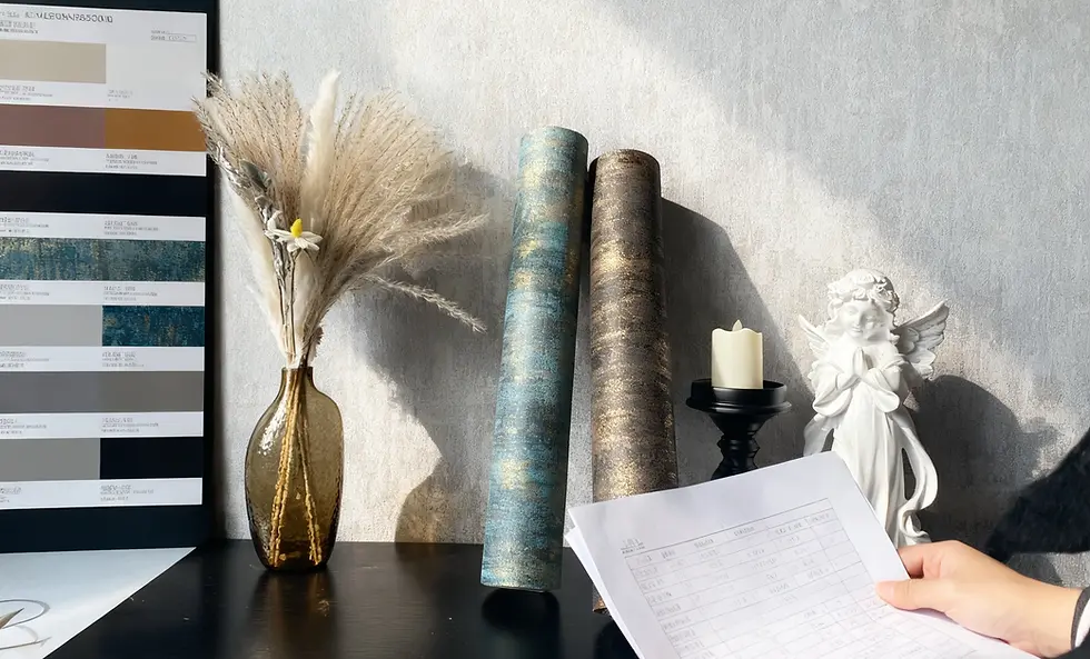 A hand holds a paper with text near a table with a vase of pampas grass, color samples, candle, angel statue, and two wallpaper rolls.