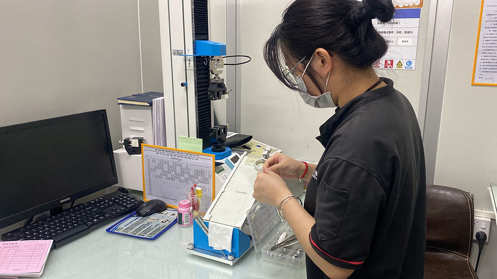 Woman in a lab coat examines samples at a desk with a computer and blue lab equipment. She wears goggles and a mask, focusing intently.