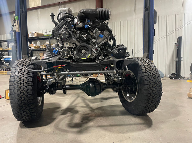 Classic Ford Bronco chassis with Ford Coyote engine, showing detailed suspension and rugged tires in a workshop setting.