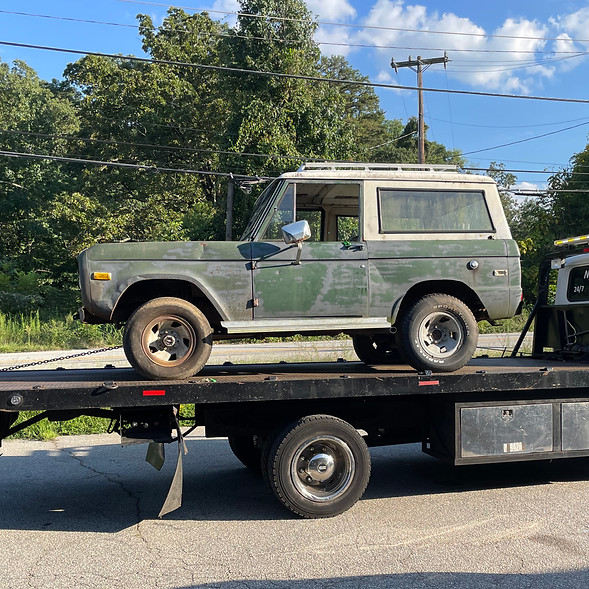 Weathered green classic Ford Bronco with a white top, loaded on a flatbed truck for transport, with trees and blue sky in the background.