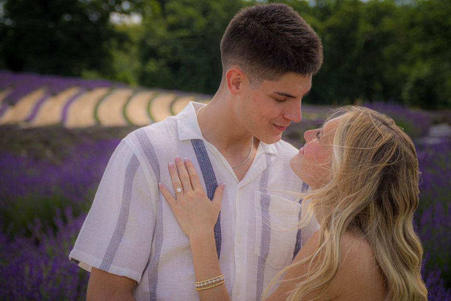 A young couple sharing a tender moment, while displaying a new engagement ring, in a lavender field in Williamston, Michigan. Wedding photos and engagement photos by 4Everscope Media (Michigan wedding photographer).