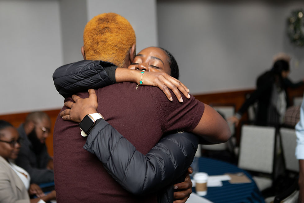 Two people hugging warmly in a conference room, one wearing a smartwatch. Others are seated in the background with coffee cups visible.