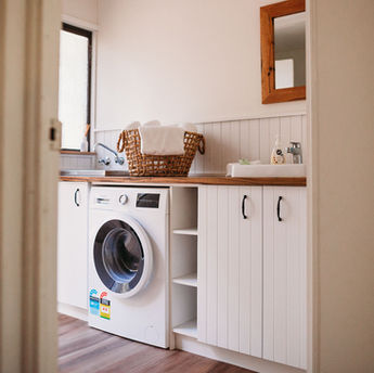Timber cottage laundry with washing machine, washing basket, sink and cupboards.