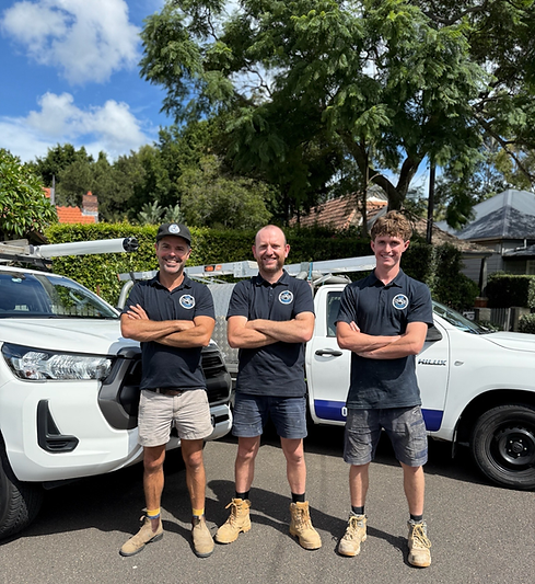 A photograph taken outdoors of three Flow Plumbing staff members standing in front of two utes, weaing brandded tshirts, shorts and work boots. They are standing with the arms crossed, smiling at the camera.