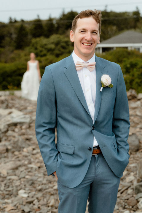 A groom smiles at the camera as a bride walks up behind him in southern Maine.