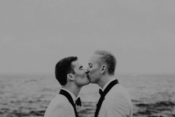 Black and white image of a gay couple kissing with water behind them.