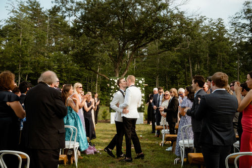 A gay couple kissing in the middle of the ceremony aisle while the guests look on and clap during a wedding near Sebago Lake, Maine.