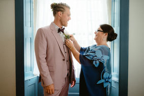 A woman helping attach a boutonniere to a groom in front of a large window in New Hampshire.