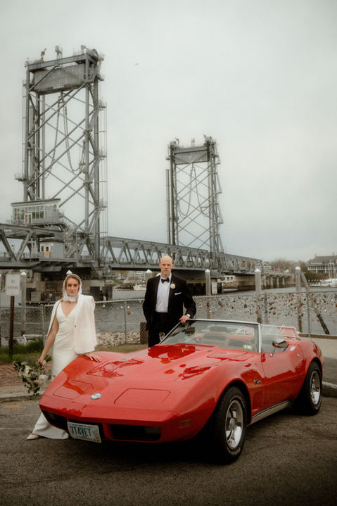 A bride and groom standing behind a classic red Corvette by the river in Portsmouth, New Hampshire before a wedding.