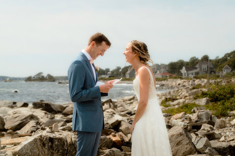 A bride and groom laughing as they read out of books on a rocky coastline in southern Maine.