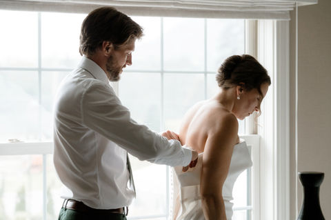 Groom helping his bride button up her dress in a hotel room in New Hampshire.