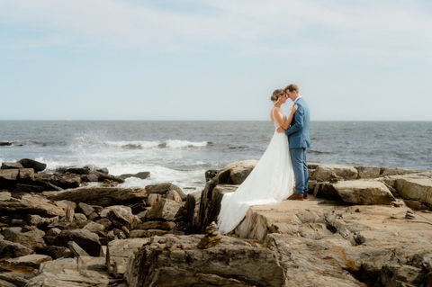 A bride and groom standing forehead to forehead as waves crash behind them on the rocky coastline in Maine.