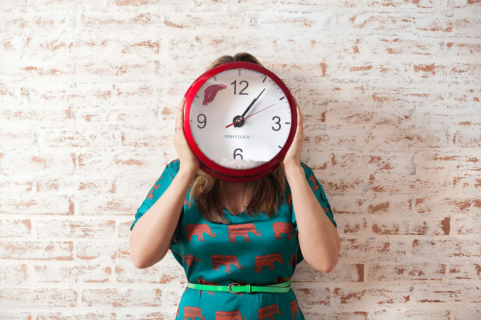 A person holds a large red wall clock in front of their face while standing against a white brick wall, representing time and deadlines.