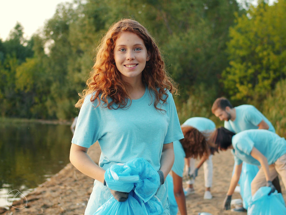A smiling young woman wearing gloves holds a blue trash bag while standing on a riverbank, with a group of volunteers in the background picking up litter along the shoreline.
