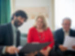 Three people sitting and reviewing documents; a man in a dark jacket, a woman in red, and an older man. They appear focused and content.