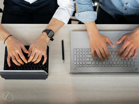 Overhead view of two people typing on separate laptops at a shared desk, with a pen placed between them, suggesting collaboration or teamwork.