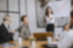A woman presents to colleagues in a meeting room, pointing to a whiteboard with charts while team members listen and take notes.
