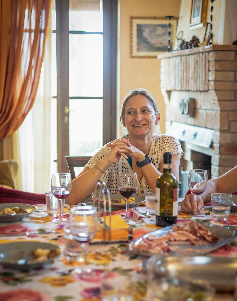 Woman smiling at dining table with wine, food, and a floral tablecloth. Bright room with a brick fireplace and framed pictures. Cozy mood.