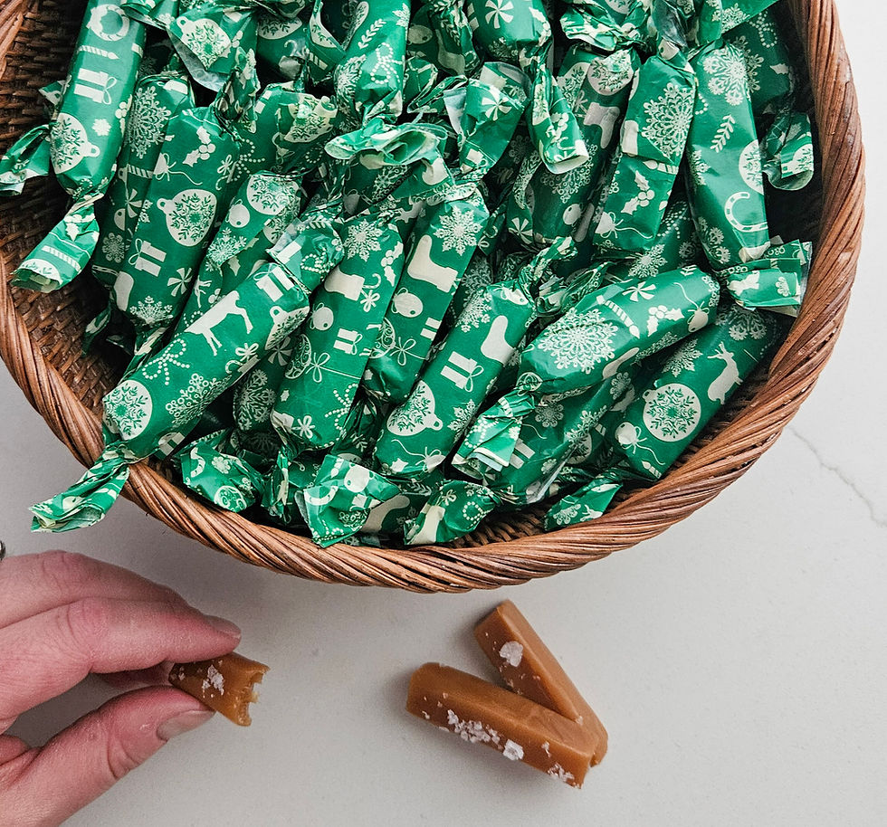 A basket full of caramels wrapped in green wrappers with Christmas designs on them, there is a hand holding a half-eaten caramel and 2 unwrapped caramels sitting on the counter