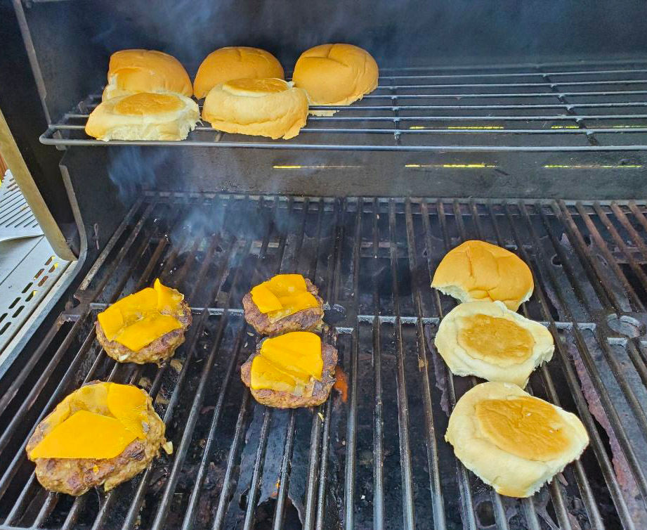 A grill with burgers and cheese and buns toasting.
